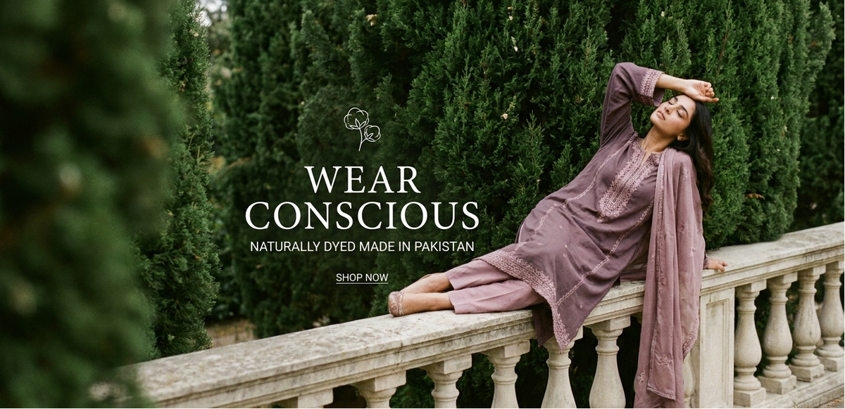 A woman reclines on a stone balustrade in front of tall green cypress trees, wearing a mauve-colored traditional outfit. The image includes the text 'WEAR CONSCIOUS NATURALLY DYED MADE IN PAKISTAN' and a 'SHOP NOW' callout.